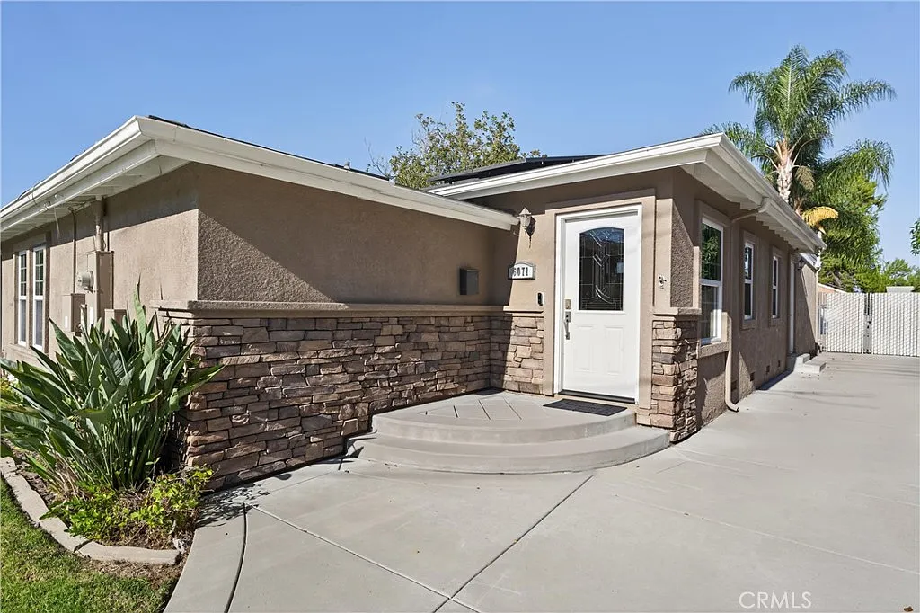 Single-story California house with beige stucco, stone accents, white door, plants, and palm trees.