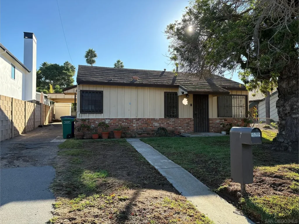 Single-story California house with beige siding, brick accents, front lawn, tree, and driveway.